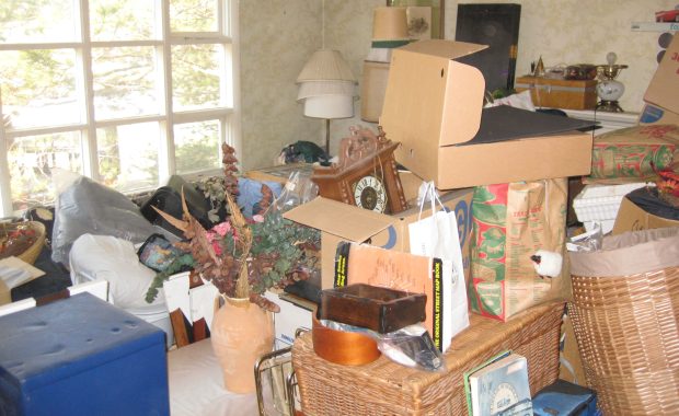 Cluttered living room filled with boxes, bags, baskets, and miscellaneous household items blocking furniture and surfaces