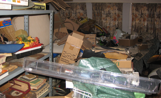 Cluttered living room filled with boxes, bags, baskets, and miscellaneous household items blocking furniture and surfaces