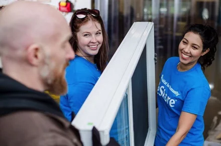 Two smiling women in blue shirts carrying a large window frame while a man in the foreground talks to them