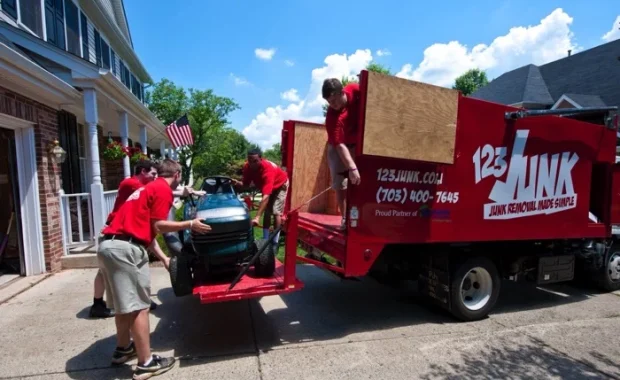 Three workers in red shirts loading a riding lawn mower onto the back of a red 123JUNK truck parked in a driveway