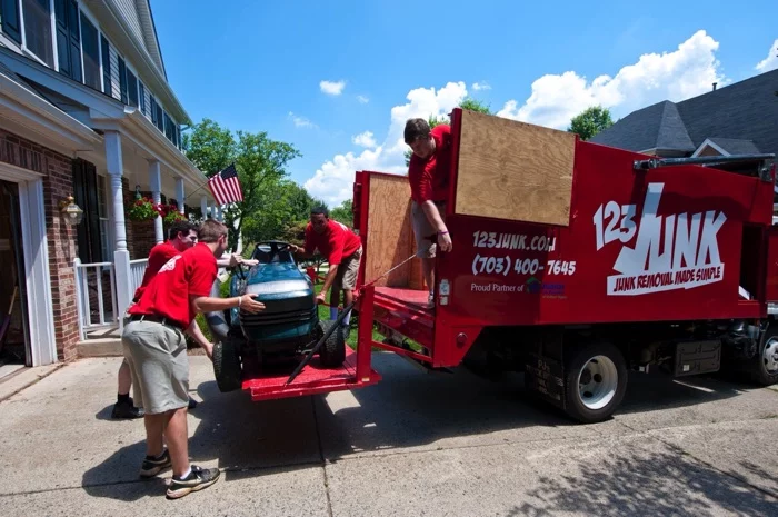 Three workers in red shirts loading a riding lawn mower onto the back of a red 123JUNK truck parked in a driveway