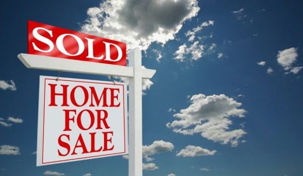 A real estate sign showing Home For Sale with a red Sold sticker on top stands against a blue sky with clouds
