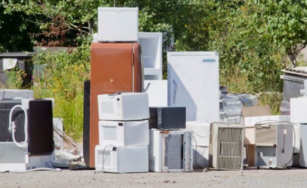 Outdoor scene showing a pile of discarded appliances including refrigerators, microwaves, and air conditioning units