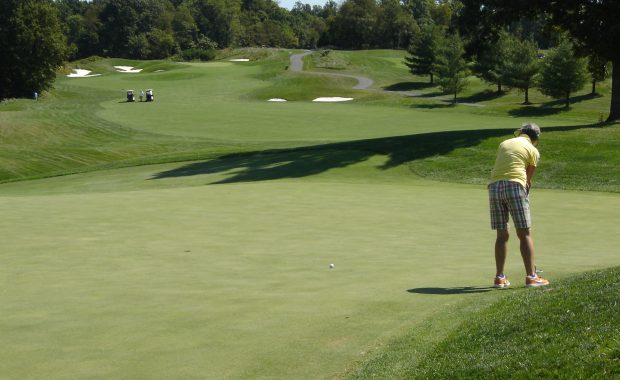 A golfer wearing a yellow shirt and plaid shorts putting on a green with sand traps and golf carts in the background