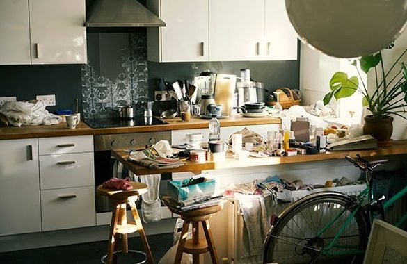 Cluttered kitchen with countertops and stools covered in various household items, and a bicycle partially visible in the foreground