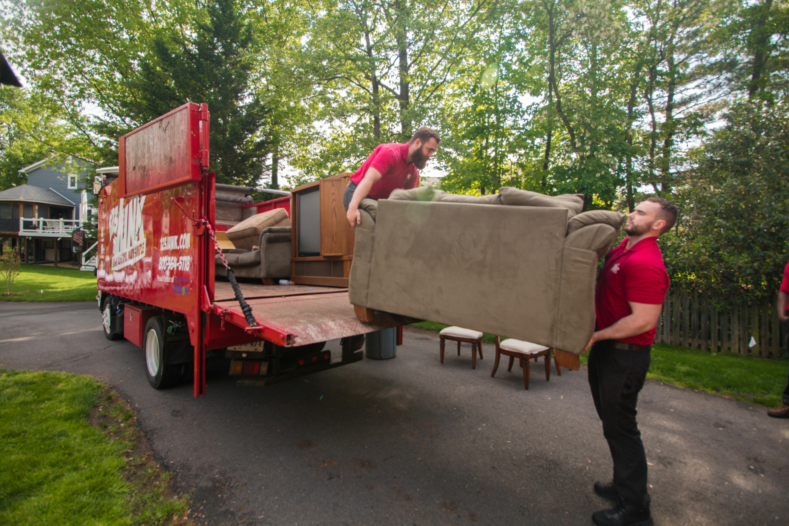 Two junk removal workers in red shirts lifting a large couch into the back of a red 123JUNK truck