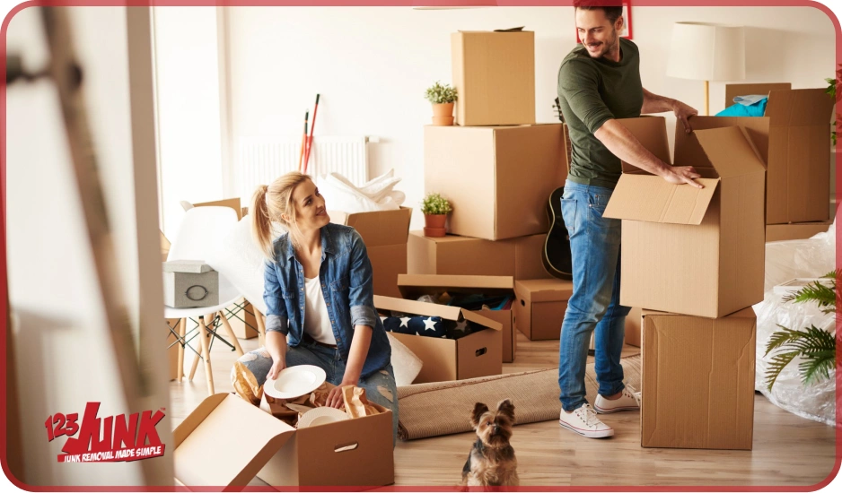 A couple packing household items into cardboard boxes with a small dog nearby and stacks of boxes in the background