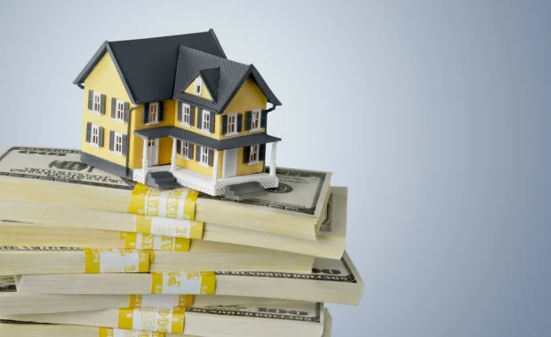 Model of a yellow house placed on top of several stacks of hundred-dollar bills against a blue background