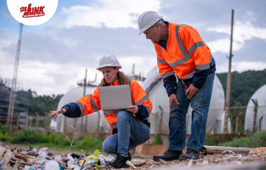 Two workers in safety gear inspect litter at an industrial site, with one holding a laptop