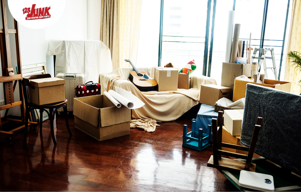 Living room filled with moving boxes, a guitar, and assorted household items stacked and covered with sheets